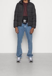 Person som bär en svart puffjacka, vinröd skjorta, ljusblå jeans, vita sneakers och en svart Adidas midjeväska med vitt logo.