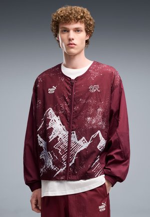 Joven con cabello rizado que lleva una chaqueta y pantalones Puma granate con diseños blancos de montañas, de pie frente a un fondo gris.