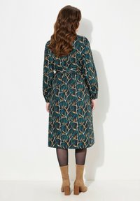 Abito lungo a maniche, di colore scuro, con una stampa floreale in teal, oro e crema, stretto in vita da una cintura coordinata. Indossato con stivali color cammello fino al ginocchio.