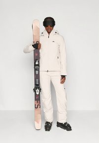 Smučarska oprema, ki vključuje svetlo bež vodoodporno jakno in hlače, črne rokavice ter črno čelado. Drži temne smuči z vezmi.