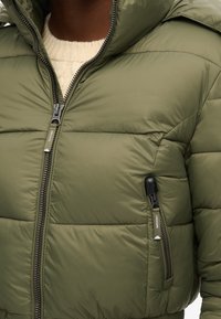 Zielona oliwkowa kurtka ocieplana z wysokim kołnierzem, zamek błyskawiczny z przodu i fakturowanym wykończeniem. Posiada boczne kieszenie oraz kontrastowe detale zamka.