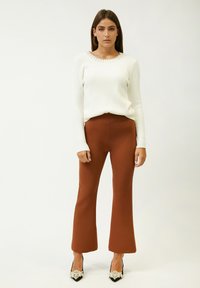 Witte geribbelde trui met een textuurkraag, gecombineerd met bruine flared broek en zwarte hakken met parelaccenten. Model staat tegen een effen achtergrond.