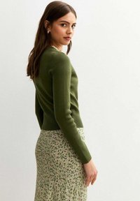 Vrouw met lang haar, naar de zijkant gekeerd, in een groene aansluitende trui en een rok met bloemmotief, tegen een lichte achtergrond.