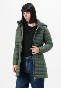 Vrouw in een open groene gewatteerde jas over een zwart shirt en blauwe jeans, staand tegen een effen witte achtergrond.