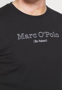 Camiseta negra con texto blanco "Marc O’Polo (Re-Nature)" llevada por una persona, mostrando el cuello y la parte superior del pecho.
