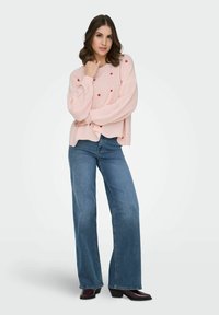 Femme debout portant un pull rose clair avec des motifs de cœurs rouges, un jean bleu à jambes larges et des bottines foncées, sur un fond blanc.