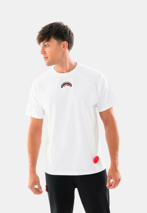 Giovane uomo con capelli corti e scuri guarda a sinistra, indossa una maglietta bianca con un piccolo disegno di bocca di squalo sul petto e pantaloni neri.