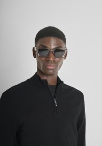 Rechthoekige zonnebrillen met een gradient donker glazen, heldere geometrische monturen. Model draagt een zwarte zip-up trui, tegen een lichte achtergrond.