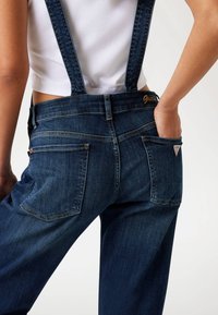 Jeans-Latzhose mit verstellbaren dunkelblauen Trägern, Metallbeschlägen und Gesäßtaschen; gesticktes "Guess"-Logo am Bund.