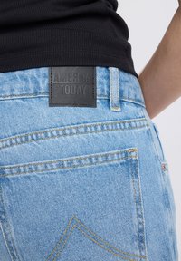 Close-up of light blue denim jeans back pocket with black leather brand label "AMERICA TODAY" and part of a black shirt above.
