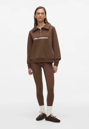 Kvinde står iført en brun lynlåstrøje med teksten "KARL LAGERFELD", matchende brune leggings, hvide sokker og brune sko.