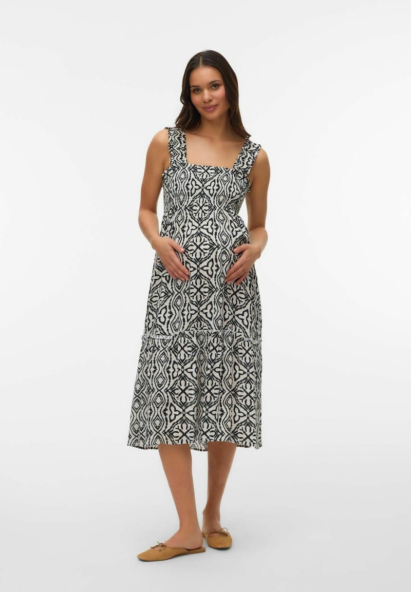 Femme enceinte portant une robe sans manches à motif noir et blanc, tenant son ventre, debout pieds nus devant un fond blanc uni.