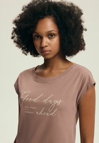 Brązowa koszulka z krótkim rękawem z białym napisem: "Dobre dni są wokół... jest pięknie. przed nami." Miękka, swobodna tkanina.