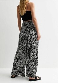 Femme portant un crop top noir à bretelles spaghetti et un pantalon large noir avec un motif abstrait blanc, se tenant devant un fond uni.