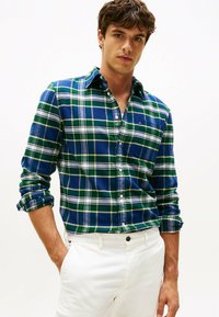 Camisa de franela a cuadros azules y verdes con líneas blancas, con cuello abotonado, mangas enrolladas, superpuesta sobre pantalones blancos con un botón negro.