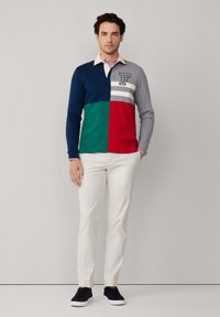 Farbblockiertes Langarm-Polohemd in Marineblau, Grün, Rot und Grau mit weißen Streifen und Logo. Kombination mit cremefarbenen Hosen und schwarzen Sneakers.