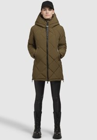 Gesteppten olivgrünen Parka mit hohem Kragen, Frontreißverschluss und seitlichen Taschen. Kombiniert mit schwarzen Leggings und kniehohen Schnürstiefeln.