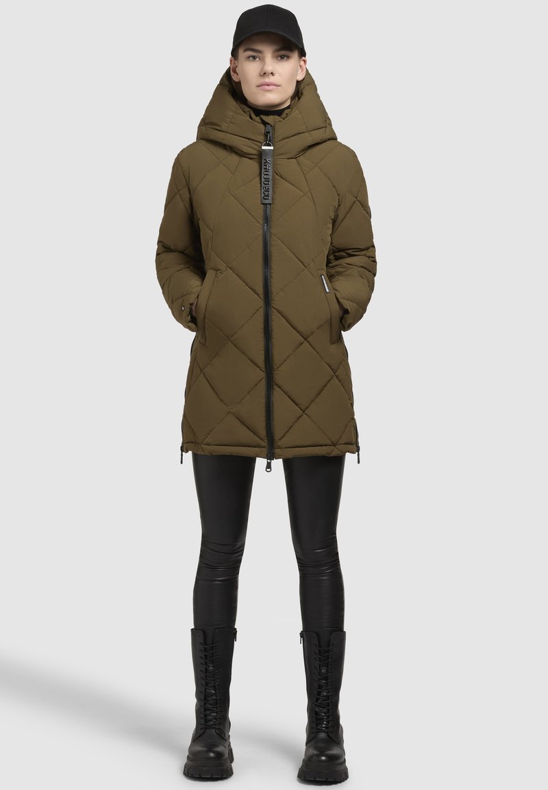 Parka verde oliva trapuntata con colletto alto, zip frontale e tasche laterali. Abbinata a leggings neri e stivali neri alti fino al ginocchio con lacci.