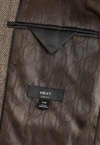 Doublure de veste à motif marron avec un rabat de poche en tissu noir et des étiquettes portant la mention "NEXT SLIM FIT" et "40R FABRIQUÉ AU BANGLADESH".