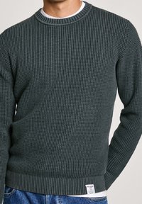 Tmavě zelený texturovaný pletený svetr s žebrovaným límcem a lemem, nošený přes bílou spodní košili a v kombinaci s modrými džínami.