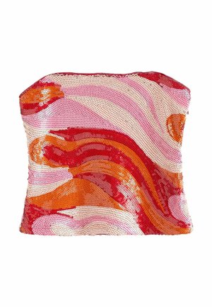 Jeune femme aux cheveux blonds ondulés portant un haut bustier rouge, rose et orange, un jean large bleu clair et des sandales, debout.
