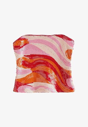 Ung kvinde med bølget blondt hår iført en rød, pink og orange stropløs top, lyseblå vidde jeans og sandaler, stående.