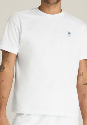 Mann trägt ein schlichtes weißes T-Shirt mit kurzen Ärmeln, auf der linken Brust ein kleines Wilson Sporting Goods-Logo und sichtbare Tätowierungen auf dem Arm.