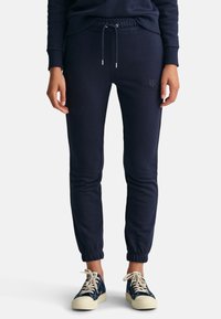 Marineblå joggebukser med elastiske mansjetter og snøring i livet. Mykt stoff med en subtil logodetalj på venstre lår. Kombinert med marineblå joggesko.