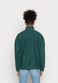 Sweatshirt verde com gola alta, tecido texturizado, ombros caídos e punhos canelados, usado com jeans azul claro.