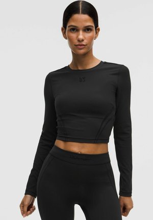 Frau trägt schwarzen langärmligen Crop-Top und hoch taillierte Leggings, steht vor einem schlichten hellen Hintergrund.