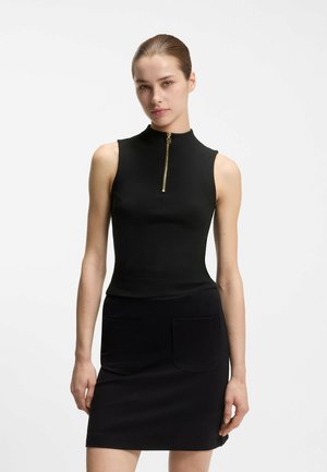 Kvinde iført en sort ærmeløs kjole med en guld lynlås foran og to lommer, stående foran en simpel hvid baggrund.