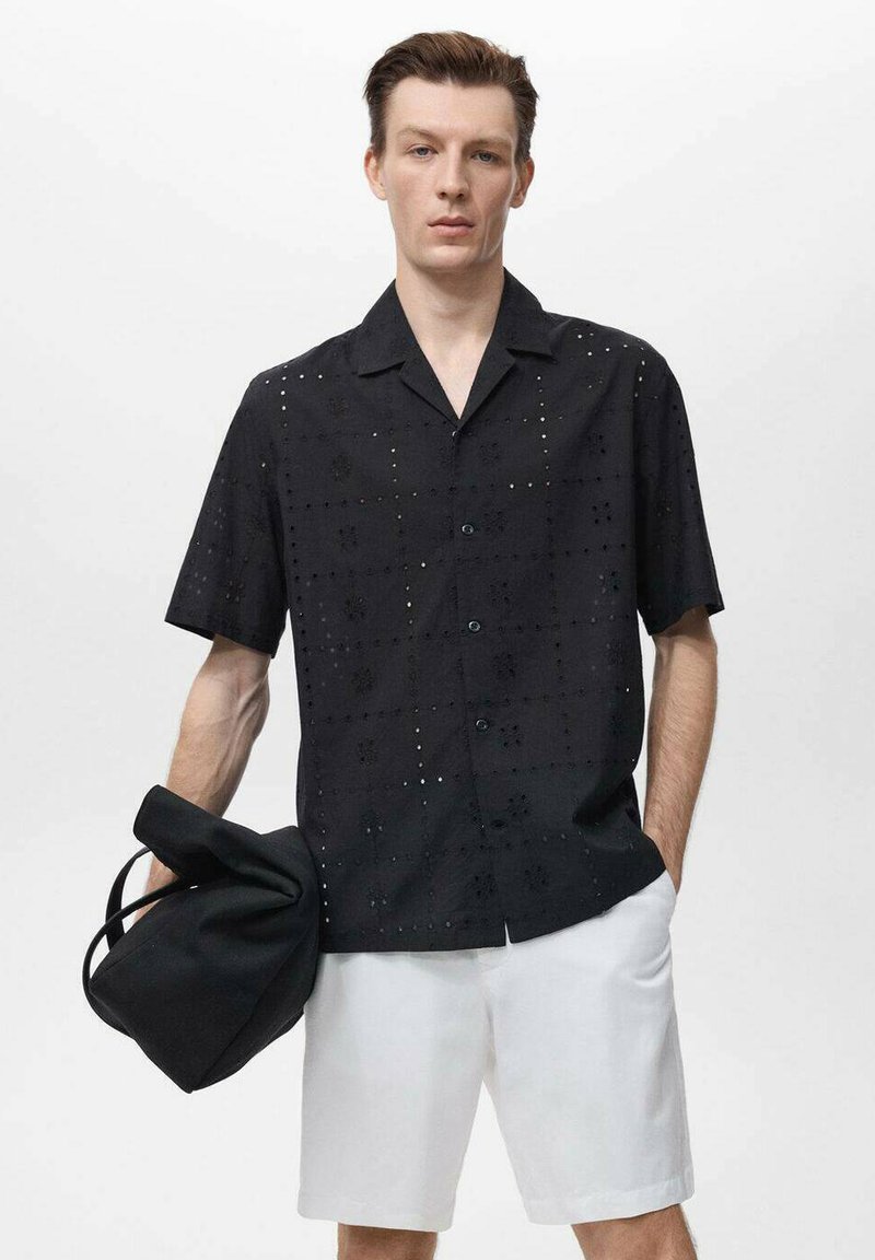 Korte mouwen zwarte shirt met gestructureerde bloemvormige uitsnijdingen, knoopsluiting aan de voorkant en een relaxte pasvorm, gecombineerd met witte shorts en een zwarte tas.