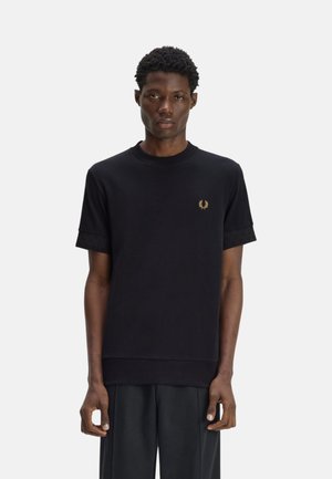 Hombre con cabello corto y rizado que lleva una camiseta negra de manga corta con un pequeño logo de laurel dorado en el pecho izquierdo y pantalones oscuros.