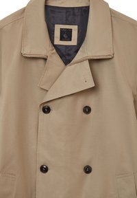 Trench coat beige doppiopetto con colletto strutturato, dotato di bottoni neri, dettagli con zip e una texture liscia. Fodera interna grigio scuro.