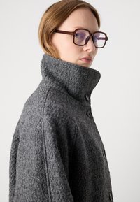 Grå uldfrakke med struktureret overflade, høj krave og sorte knapper. Bæres med firkantede briller, som fremhæver frakkens sideprofil.