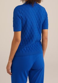 Kvinna i en blå kortärmad texturerad stickad topp och matchande blå byxor, sedd från ryggen mot en neutral bakgrund.