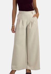 Pantalon à jambes larges en tissu beige clair avec plis, taille haute et poches latérales. Texture lisse et lignes épurées sur l'ensemble.
