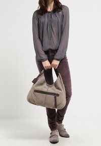 Šedá taška hobo s hladkým, měkkým povrchem, která má přední zipovou kapsu a hnědou akcentovou šňůru. Nositelná s button-up, modrošedým topem a fialovými kalhotami.