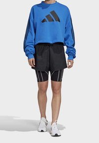 Sweat-shirt court bleu avec logo noir, assorti à un short noir porté sur des leggings noirs à rayures, et des chaussures de sport blanches.