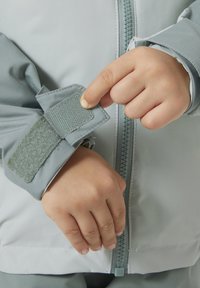 Světle šedá bunda se zapínáním na zip a úpravou manžet na suchý zip. Má hladkou texturu a zesílené švy na manžetách.