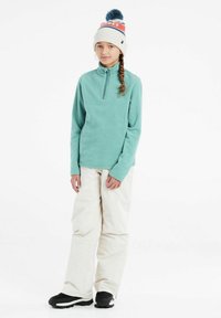 Pullover in pile color turchese con scollatura a metà cerniera, abbinato a pantaloni da neve bianchi e stivali neri. Berretto con pom pom blu e dettagli rossi.
