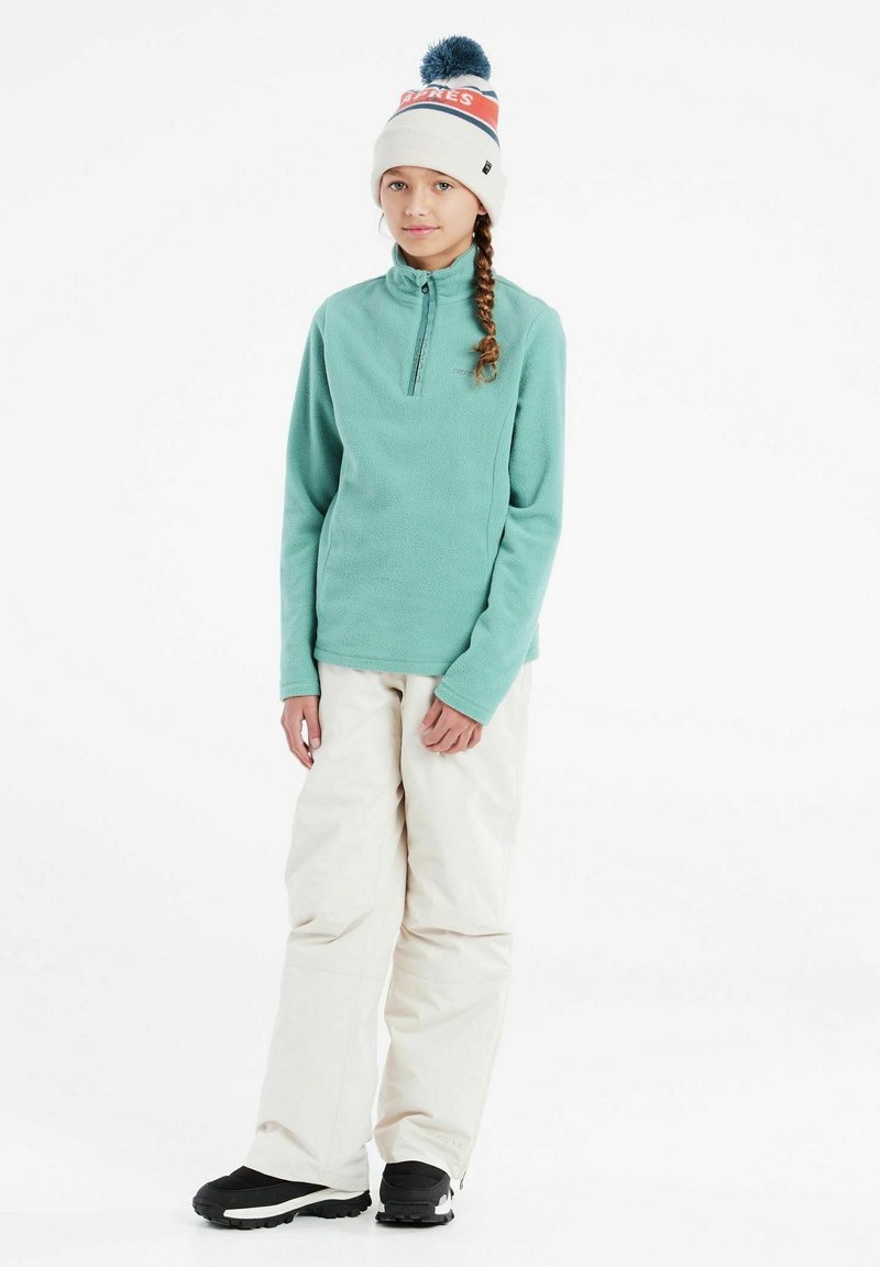 Pullover in pile color turchese con scollatura a metà cerniera, abbinato a pantaloni da neve bianchi e stivali neri. Berretto con pom pom blu e dettagli rossi.