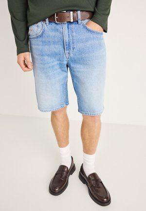 Hellblaue Jeansshorts mit ausgewaschenem Finish, einem braunen Ledergürtel, kombiniert mit weißen Socken und dunkelbraunen Loafers.