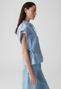 Lichtblauwe top met korte mouwen, een kraag, een borstzak en verstelbare strikjes in de taille. De stof lijkt glad met een gestructureerd ontwerp.