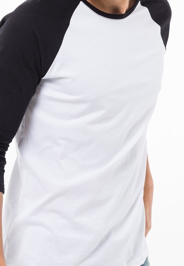 Homme portant un t-shirt de style baseball blanc avec des manches raglan noires, debout devant un fond blanc uni.