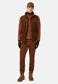 Gilet trapuntato marrone sopra un maglione a costine, abbinato a pantaloni marroni e guanti. Il modello indossa un cappello marrone coordinato e stivali marroni.