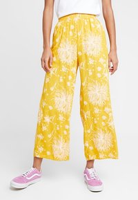 Gula blommönstradevida byxor med elastisk midja, gjorda av ett lätt material, i kombination med rosa sneakers.