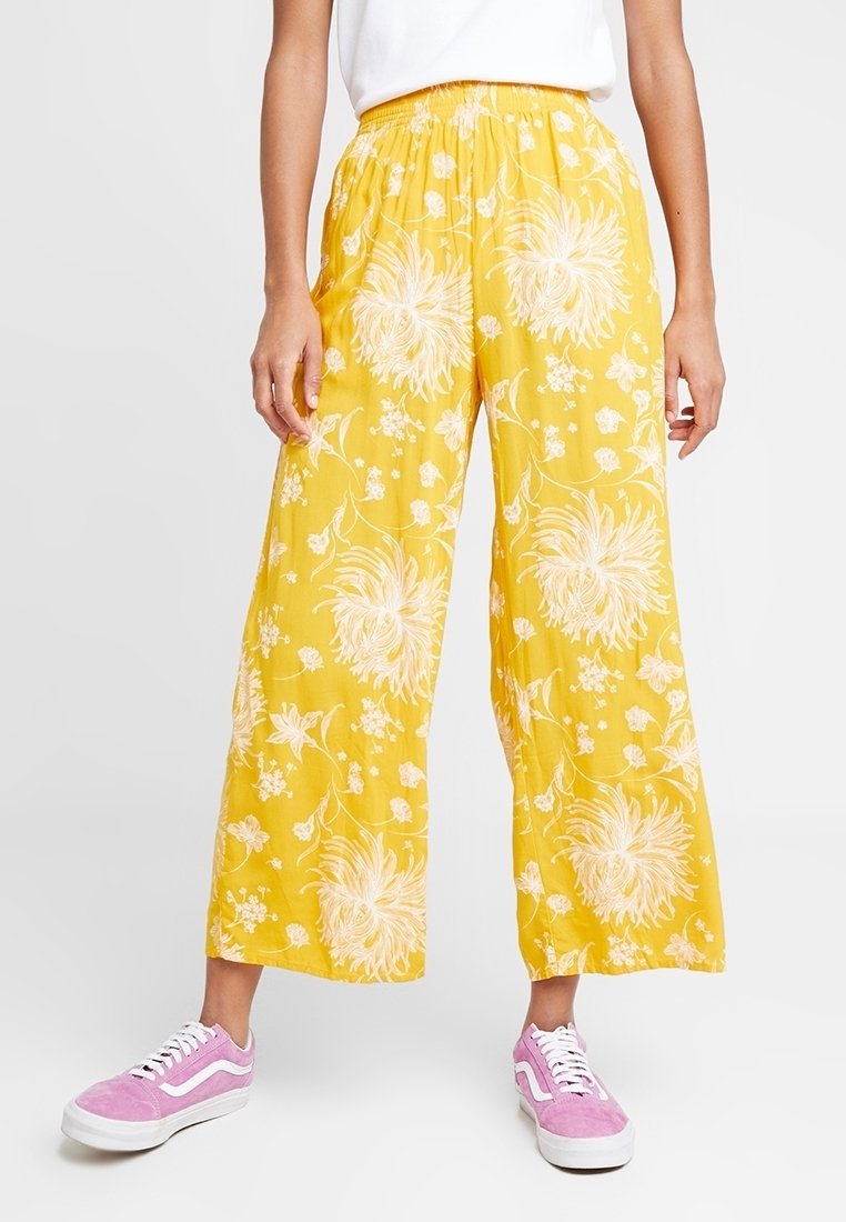 Gula blommönstradevida byxor med elastisk midja, gjorda av ett lätt material, i kombination med rosa sneakers.
