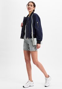 Vrouw met een glimlach, gekleed in een marineblauwe bomberjack, gestreepte denimshorts, witte sneakers en een lichtgrijze schoudertas, tegen een witte achtergrond.