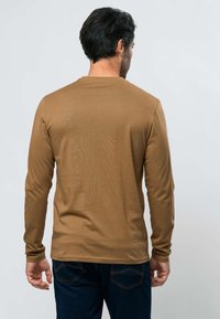 Homme portant une chemise marron à manches longues et un jean bleu foncé, debout de dos devant un fond uni gris clair.